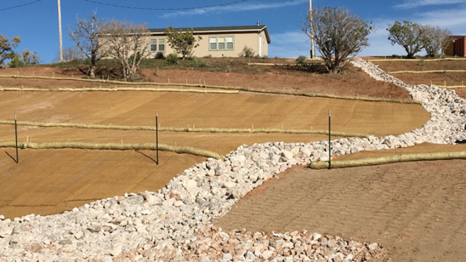 Steep slope no longer showing signs of water erosion. Instead berms in place and a large, stone filled channel for water goes up the slope leading to a home