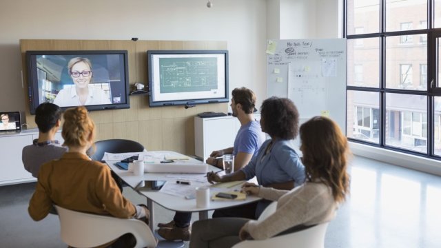 Team sitting around a table collaborating with a person virtually on a large screen in front of them.