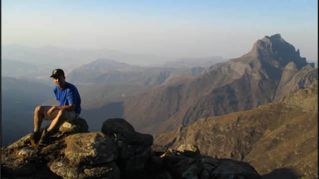 Sam Suzuki sits on a mountain top in South Africa with a vista of mountains behind him