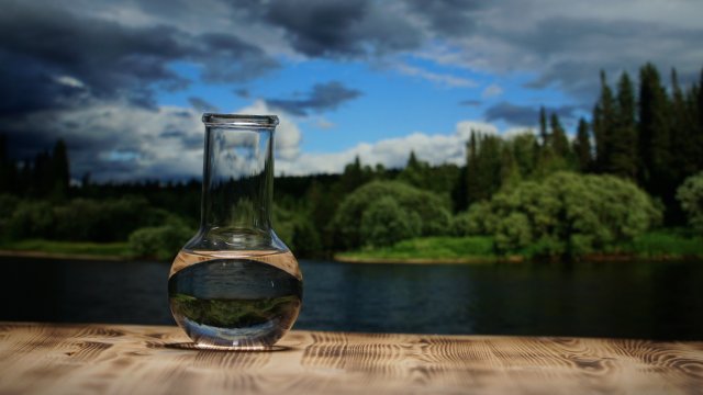 Clean water in a glass laboratory flask on wooden table on mountain river background.