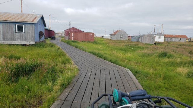 Pathway bordered by green grasses running through the rural Alaska Native Village of Kongiganak. Taken from the perspective of someone driving an ATV.