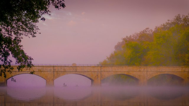 Bridge crossing river with people on water and trees along river's edge.
