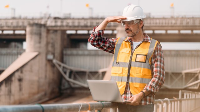 An engineer in a hardhat with a laptop looking at a treatment plant