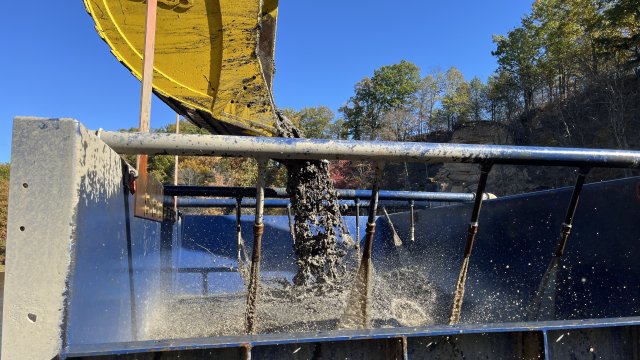 Mud pours out of a yellow excavator bucket as it hovers over a large receiving tank. 