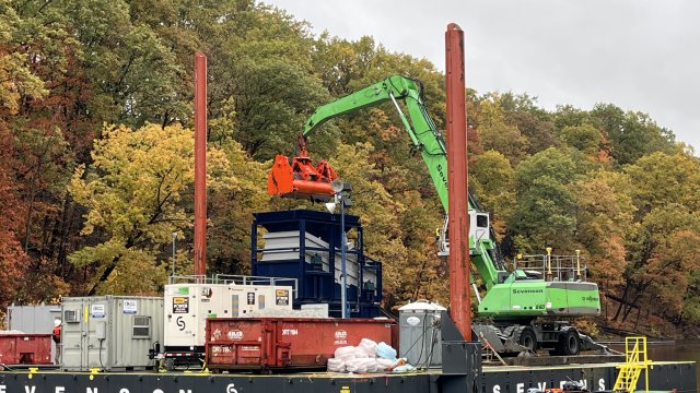 Construction equipment sit on top of a floating platform on the river with colorful trees in the background. 