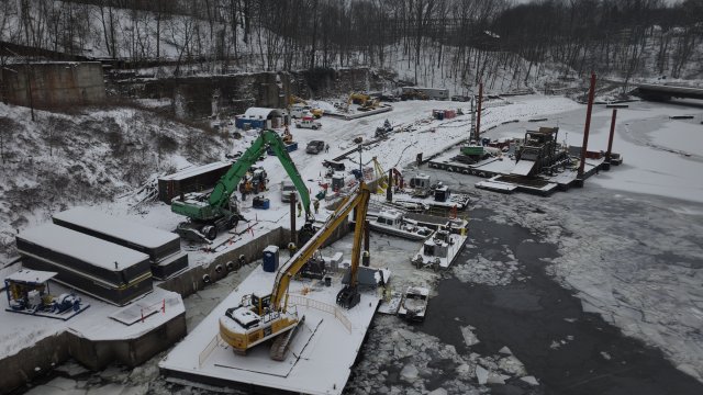 Construction equipment on floating platforms in river next to construction equipment on the shoreline. River is ice-covered and snow coats everything. 