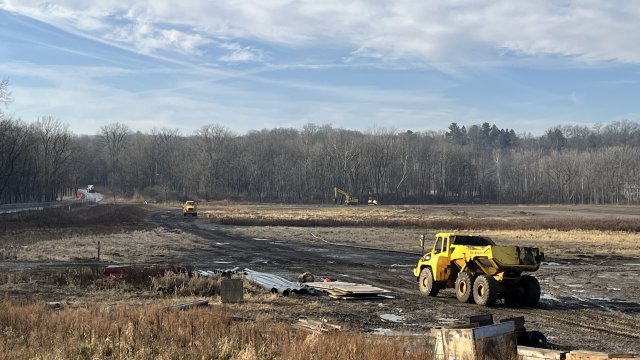 A large yellow truck drives across a field of brown soil and dried grasses. Brown, bare trees fill the background along with a blue sky and some white cloud cover. 