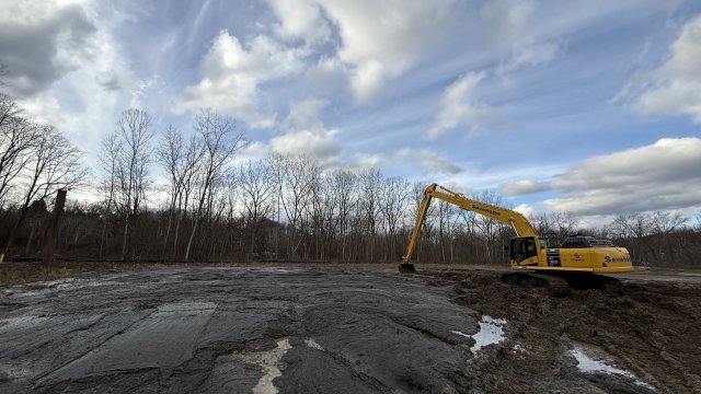 A large yellow excavator uses its long arm and clamshell bucket to spread gray-brown sediment into a flat, even layer. There are bare trees in the background and a blue sky with fluffy white clouds.