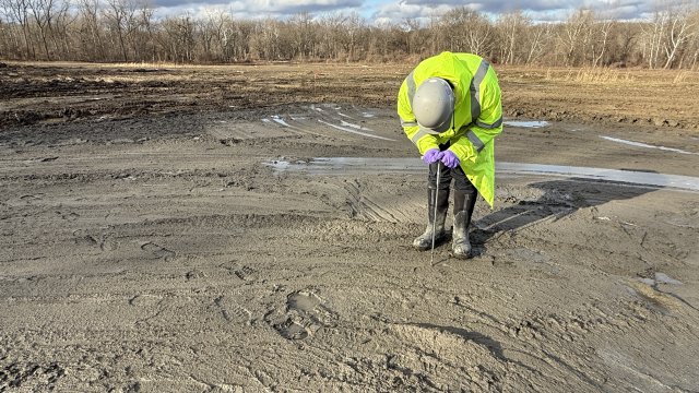 A construction crew member in a gray hard hat, purple gloves, and bright yellow jacket stands on a flat patch of gray-brown sediment and presses a long, thin metal instrument into the ground. Bare trees and a blue sky with fluffy white clouds fill the background.