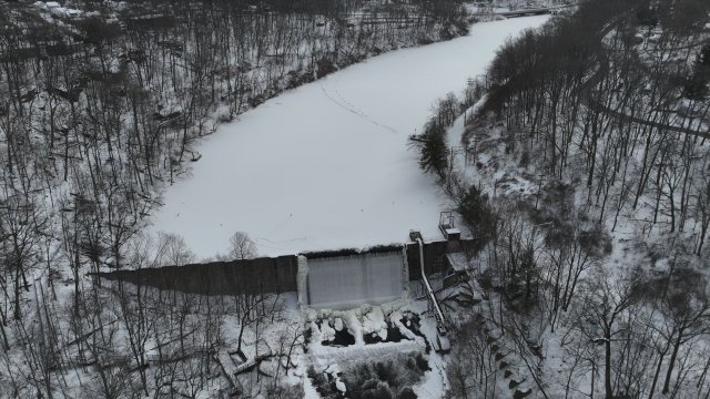 A birds-eye view of a frozen river covered in snow. A white waterfall flows over a large dam, with snow- and ice-covered boulders at the base. The area surrounding the river is forested with bare trees covered in snow.