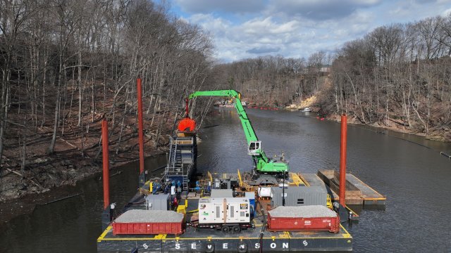 A large green crane and other dredging equipment on a black platform barge float at the surface of a river of dark water. Bare trees line the riverbanks and the blue sky is full of white clouds.