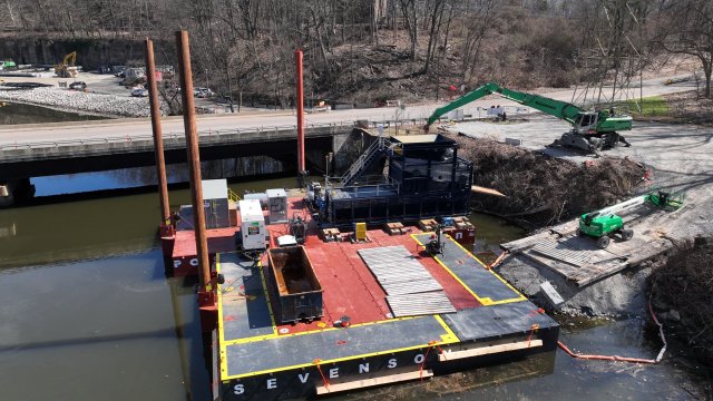 A large green crane on the bank of a river lifts a piece of metal dredging equipment onto a red and black dredging barge. The barge has tall, red poles around the perimeter that anchor it in place in the river bottom. A four-lane bridge is in the background with bare trees on the riverbank.