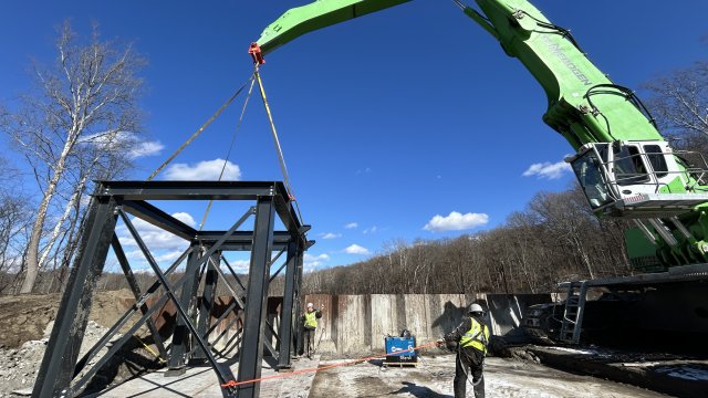 Black metal scaffolding being lifted by a large green crane while construction crew members use rope to guide the scaffolding into place on the ground. The background is filled with bright blue skies and bare trees.