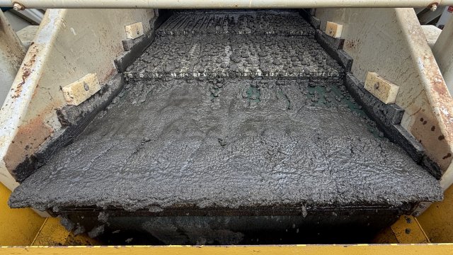 A thick dark brown slurry of sandy sediment spread across a wide, flat conveyor belt. Yellow and white steel panels frame the machinery.