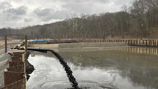 Dark muddy water pours out of a black pipe into a large basin of water. The basin walls are made of corrugated steel sheet piles. Bare trees fill the background along with a gray, cloudy sky.