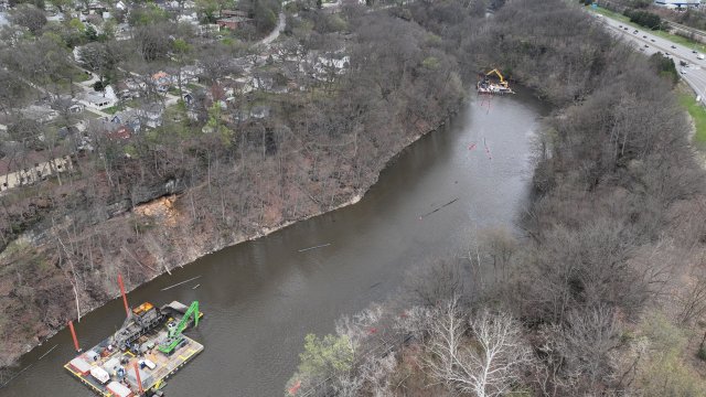 An aerial view of a wide river spans the frame. A barge with heavy construction equipment is at each end of the river, one in the bottom left of the frame and one in the top right of the frame. The river is surrounded by bare trees, suburban neighborhoods on the left, and a highway on the right.