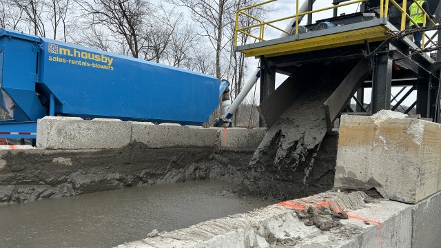 Dark gray mud pours out of a conveyor belt into a white cinderblock basin. Large yellow and blue construction equipment is in the background along with bare trees and a gray overcast sky.