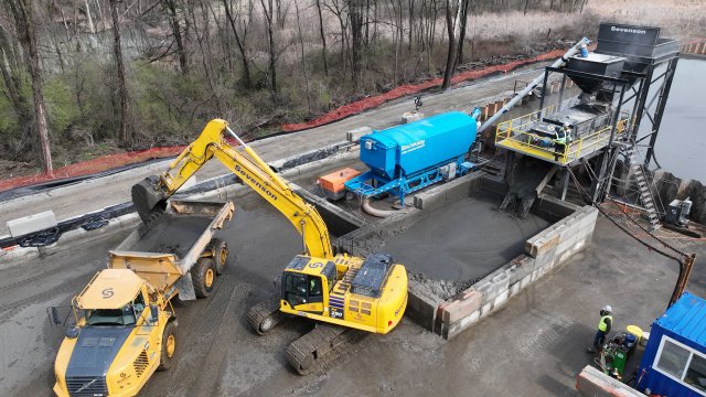 A birds-eye view of a flat, gray, bare earth construction site with large yellow and blue construction equipment. A large yellow crane scoops gray mud out of a white cinderblock basin and drops it into the bed of a large yellow hauling truck. Orange construction fencing and bare trees are in the background.