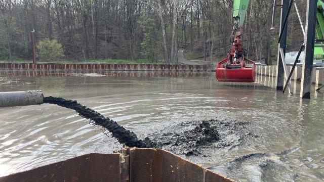 Dark brown mud spurts out of a black pipeline on the left. The mud is pouring into a large basin of brown water surrounded by rusty steel sheet pile walls. A red excavator bucket attached to a green crane can be seen in the background, along with a forest of budding trees.