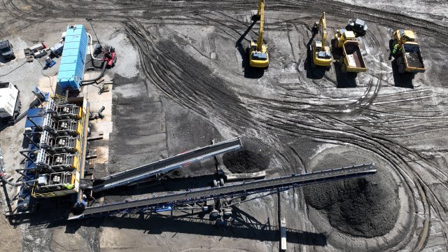 A birds-eye view of a flat, gray, bare earth construction site with two conical piles of sand and debris. Large yellow trucks and cranes are seen from above, along with a large piece of machinery with many pipes and two long conveyor belts projecting out over the piles of material.