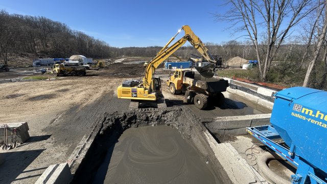 A large yellow excavator scoops grayish brown mud out of a large cinderblock basin and transfers it into the bed of a large yellow hauling truck. A piece of bright blue construction equipment is seen in the foreground, with more equipment and bare trees in the background.