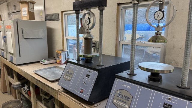 Image shows the inside of a field laboratory. The lab bench is constructed from wood, and two large compacting instruments with pressure gauges are in the foreground. A cylinder of gray mud is being compressed on the silver plate of one of the instruments.