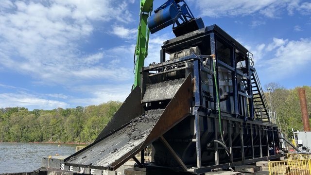 A large blue and green piece of marine construction equipment is shown floating on a river. The machinery has a large steel ramp attached to it, and black leafy muddy debris can be seen collecting in a large rusty steel basin at the base of the ramp. In the background are bright green trees and blue sky with white clouds.