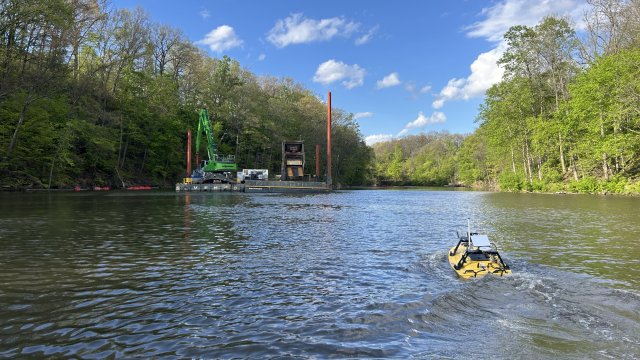 A small yellow boat approaches a large dredging barge upstream on a river. The sky is blue with white clouds, and the riverbanks are covered with bright green trees.