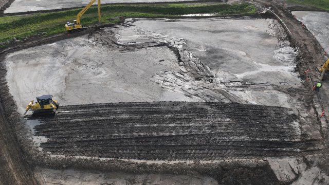 A yellow bulldozer moves across a flat expanse of gray soil. The bottom half of the photo is covered with rough textured soil and the top half is covered with smooth soil. Patches of grass are in the background with a large yellow excavator.
