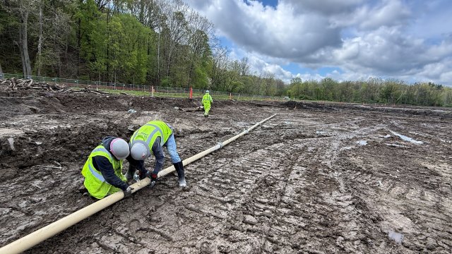 Two construction crew members in bright green safety vests and gray hard hats assemble a white pipeline. They are surrounded by bare soil. Another crew member in bright green safety gear walks in the background. The sky is blue and filled with large white clouds, and bright green trees fill the background.