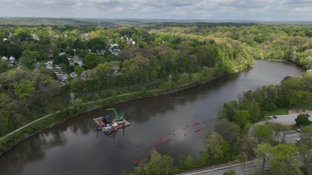 A birds-eye-view of a large river with still, dark water. On the river is a dredging barge with large green excavator equipment, and bright red buoys float on the river’s surface. A 4-lane road crosses the bottom of the photo. The river is surrounded by green trees and white houses, and the sky is full of gray fluffy clouds.