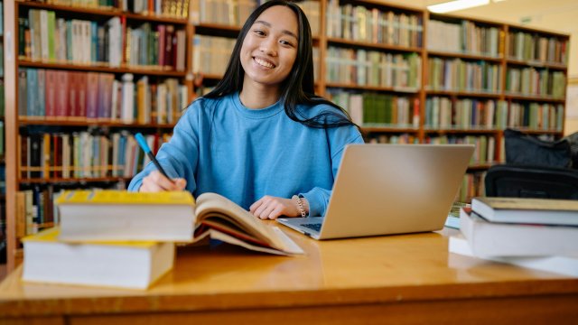 Smiling young woman studying in a library with books and laptop.