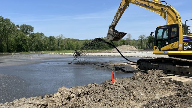 Dark sediment pours from a black pipe attached to the bucket of a large yellow excavator. Dried grayish brown earth is in the foreground, and the background is filled with green trees and clear blue skies.