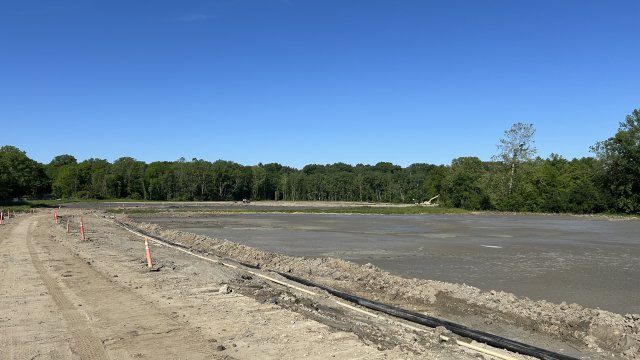  A ground-level view of a field with exposed tan soil. Green leafy trees line the field, and a blue sky is in the background.