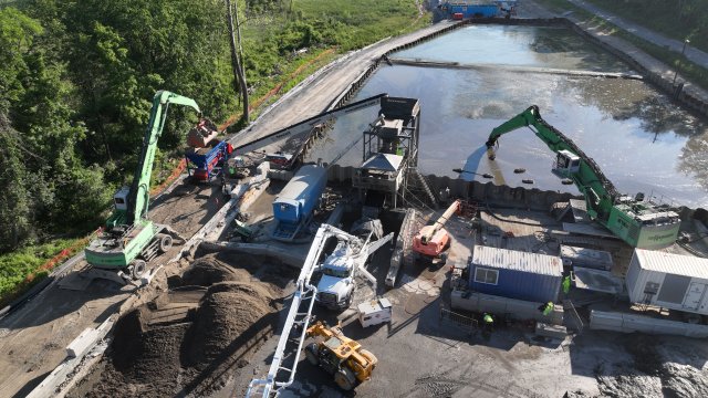 An aerial view of an active construction site with several pieces of large construction equipment in close proximity. A large basin of still water is in the background, and green trees surround the site.