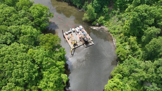 A bird’s eye view of a square dredging barge with large yellow construction equipment float on a dark river surrounded by green trees. 