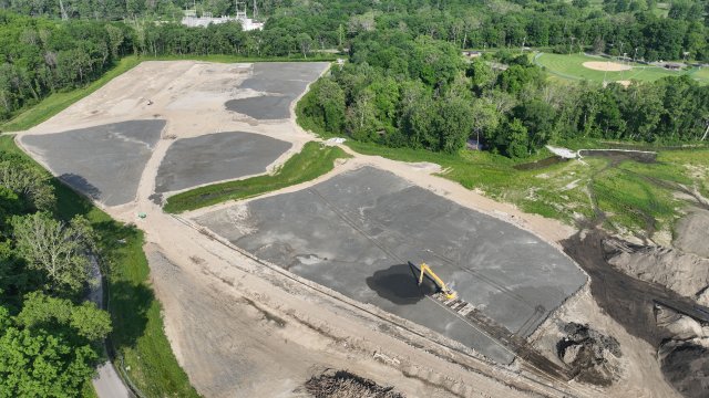 An aerial view of a large flat field. Much of the field is exposed tan soil, with patches of dark gray soil and green grass intermixed. Leafy green trees surround the field.