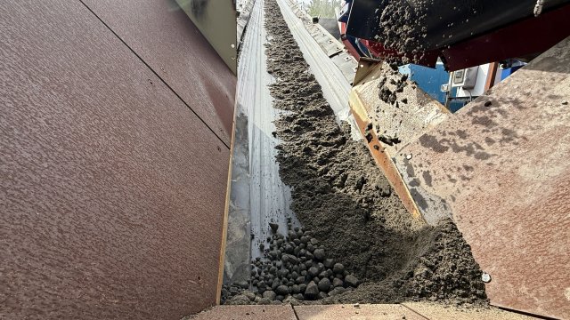 An upward view of a gray conveyor belt carrying coarse brown sand up and out of frame. The foreground is bordered by brown composite planks.