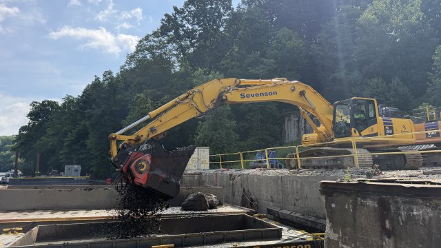 A large yellow piece of construction equipment pours shredded black organic matter into a large rectangular steel basin floating on brown water. Green trees and a blue sky with white clouds fill the background.