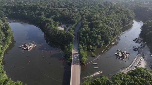 A bird’s-eye view of a wide river bisected by a 4-lane roadway. Large dredging barges float on the water on both sides of the bridge. The river is surrounded by green trees, and the sky is a hazy blue.