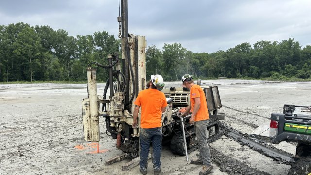 : A cream-colored piece of heavy machinery is being operated by two construction crew members in hi-visibility orange shirts and white hard hats. The crew members and equipment rest on top of a flat expanse of gray earth. Green trees and a cloudy sky fill the background.