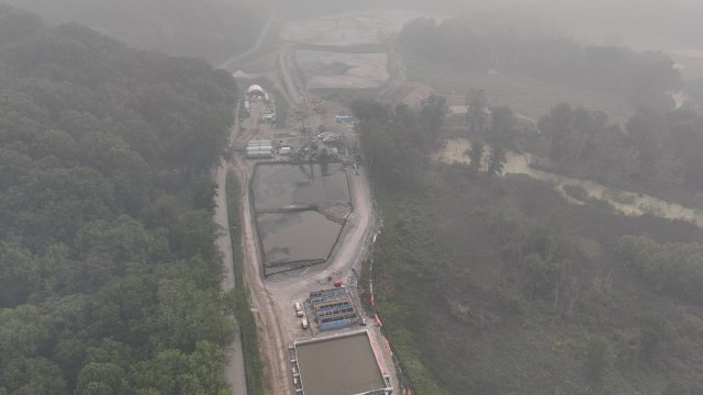 A bird’s eye view of a construction area with large equipment surrounded by green trees. Thick, hazy smoke shrouds the view.