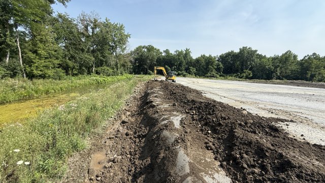 A brown, earthen berm surrounded by green vegetation on the left and light gray soil to the right. A yellow excavator is in the background, and the sky is blue with white wispy clouds.
