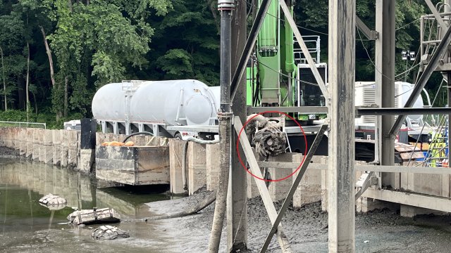 Steel scaffolding, sheet pile, and other construction equipment can be seen with brown sediment in the foreground. A large metal conical grinder can be seen between the scaffolding.