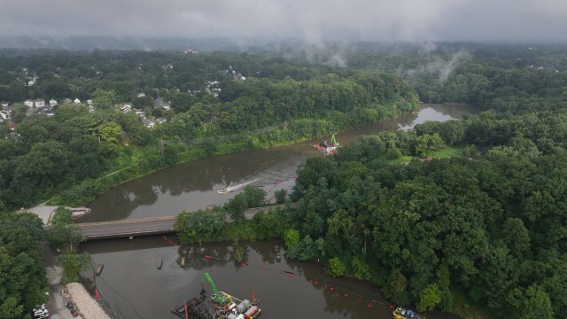 A birds-eye view of a river bend with large floating construction equipment. The river is bisected by a road bridge and bordered by green trees. The sky is gray and low-hanging clouds form mist over the trees.
