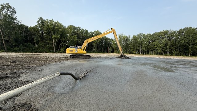 Gray, fluid mud spurts from a white pipeline onto a muddy field. A large yellow excavator scoops and levels the mud in the background. The field is surrounded by green trees and a blue sky.