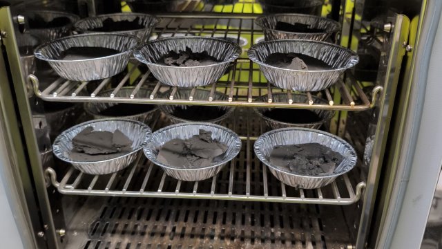 A gray metal oven with shiny, reflective interior and two metal wire racks holds small aluminum pie tins filled with dried cakes of mud.