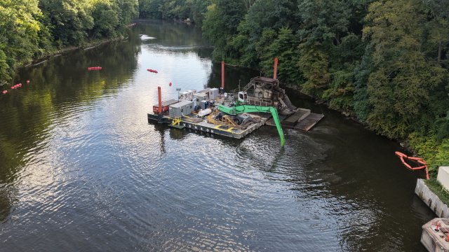 A birds-eye view of a calm river. The surface of the river is reflecting the blue sky and puffy white clouds. A barge floats on the river and a large green excavator arm extends below the surface of the water. Green trees are on the banks of the river. 