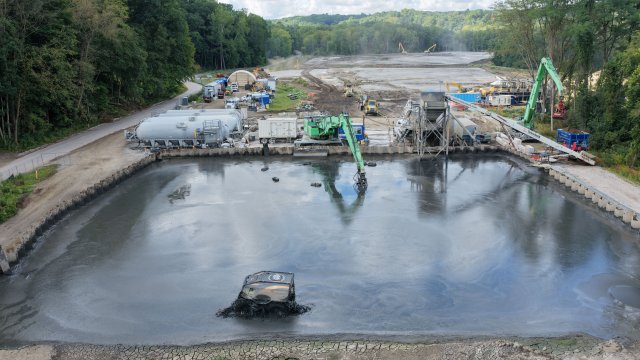 An aerial view of a construction site with large basin with sheet pile walls filled with watery sediment. A small vehicle drives across the surface of the basin stirring up the sediment. Large green excavators are in the background along with green trees.