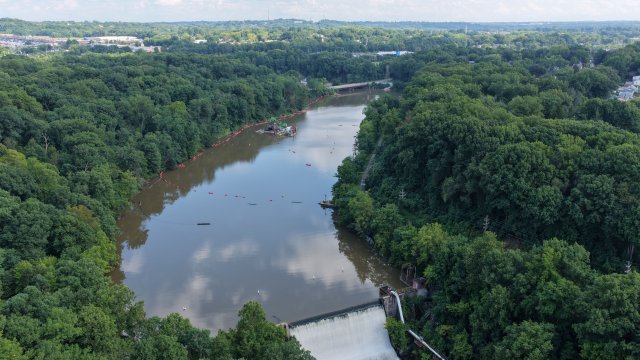 A birds-eye view of a reservoir and a large dam with whitewater flowing over. The reservoir surface is smooth and still, reflecting the blue sky with puffy white clouds. Green lush trees surround the reservoir.